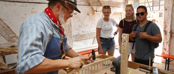 Mann bearbeitet vor drei Zuschauenden mit einem Hammer einen Holzrechen