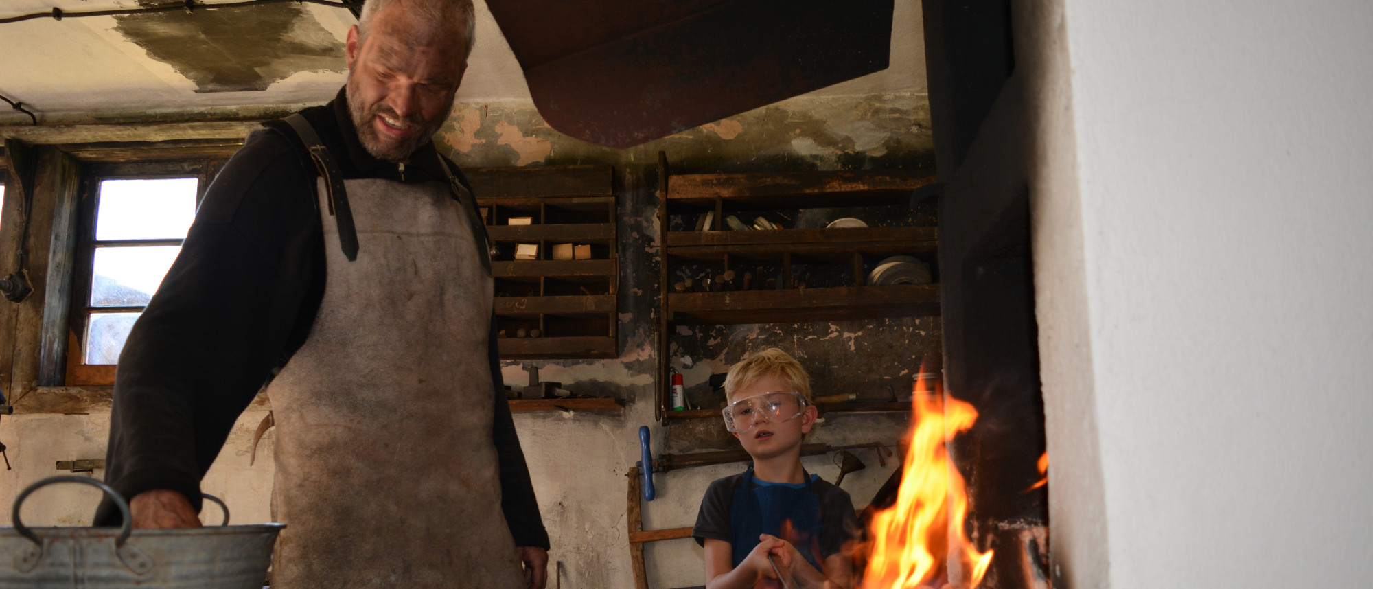 Kind schmiedet am offenen Feuer, daneben der Schmied