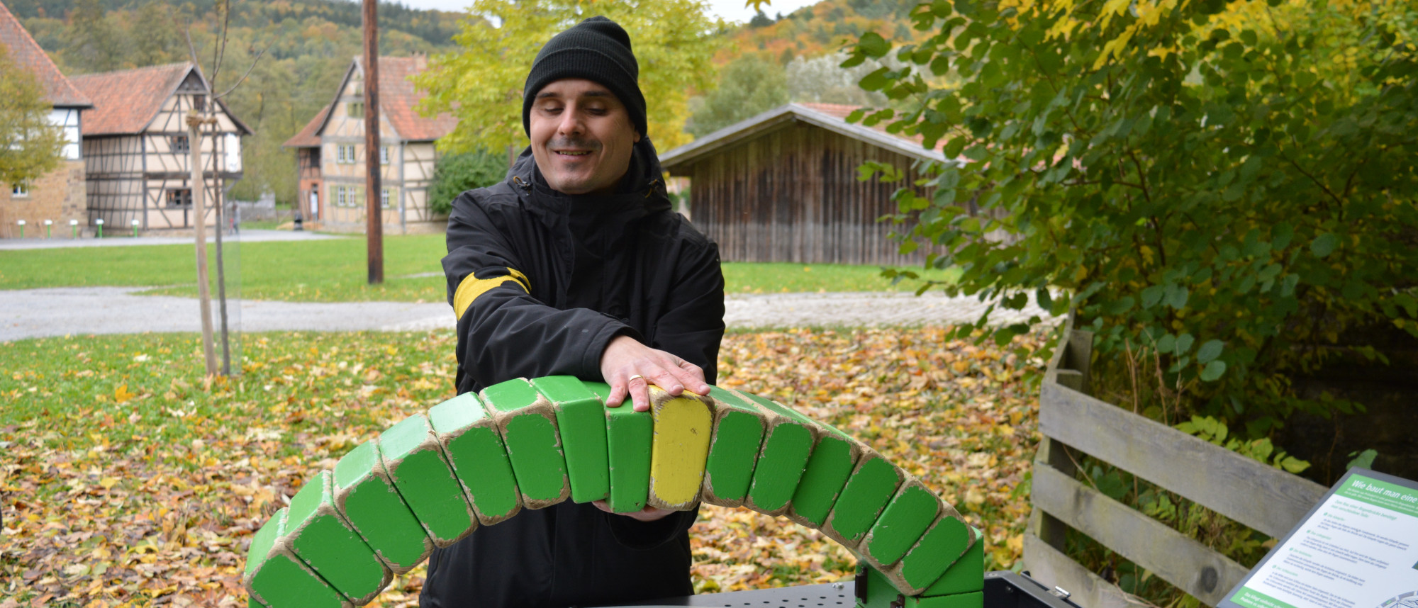 Ein Mann betastet eine kleine Brücke, dahinter Wiesen, Bäume und Fachwerkhäuser