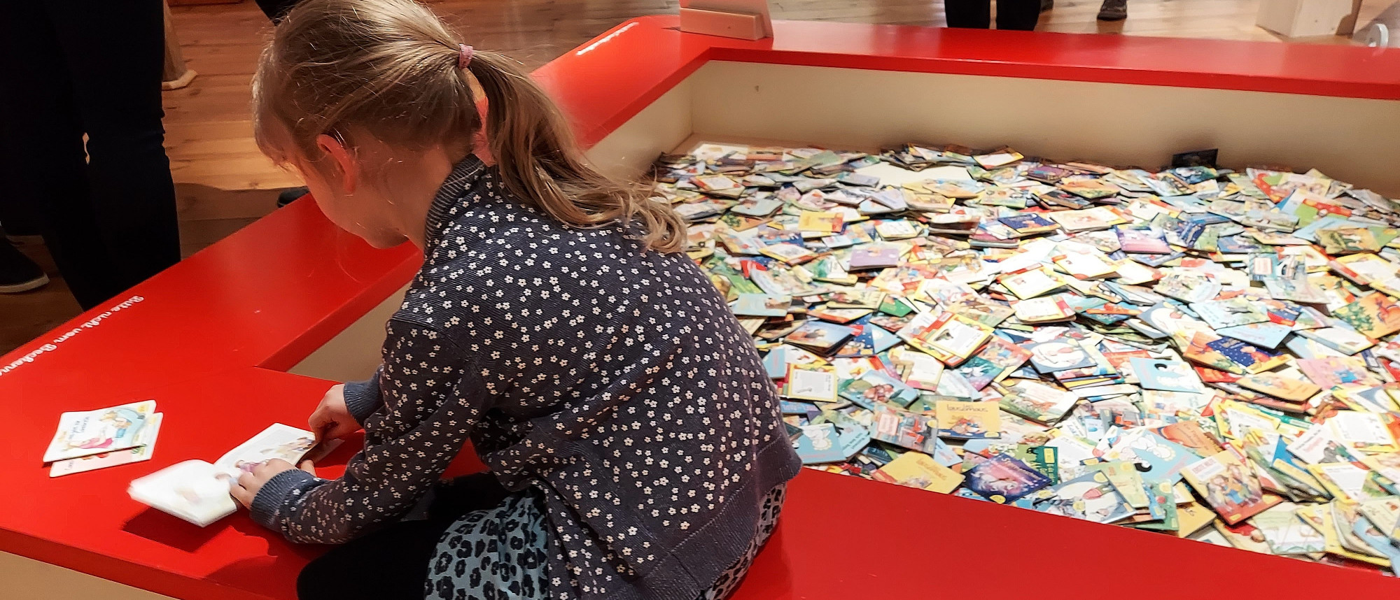 Mädchen liest in einem Buch, auf dem Rand eines Sandkastens sitzend, der mit Büchern gefüllt ist