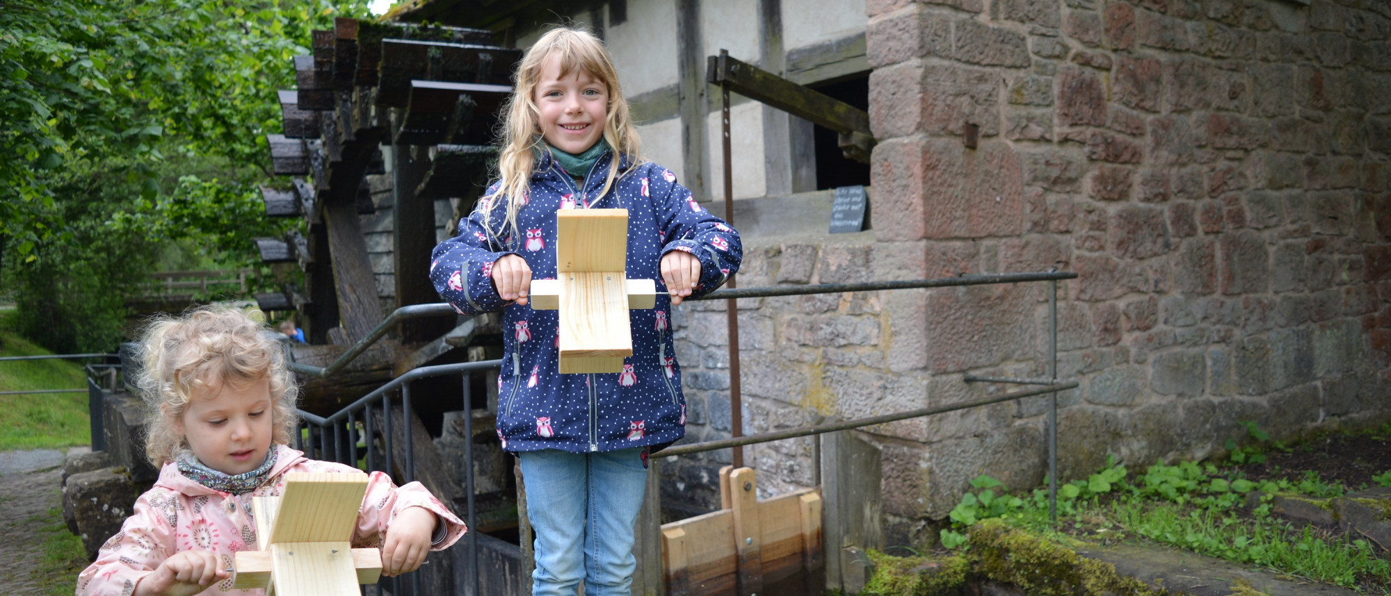 Zwei M&auml;dchen stehen vor einer M&uuml;hle und halten kleines Wasserrad aus Holz in der Hand