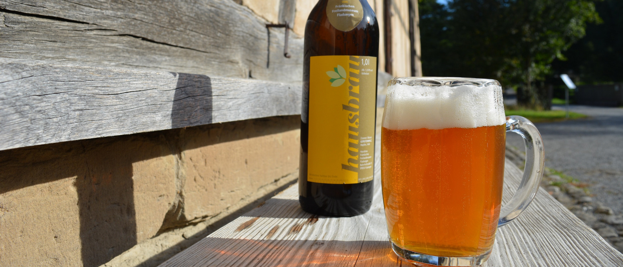 Bierflasche neben gefülltem Bierglas auf einer Holzbank vor Fachwerkhaus