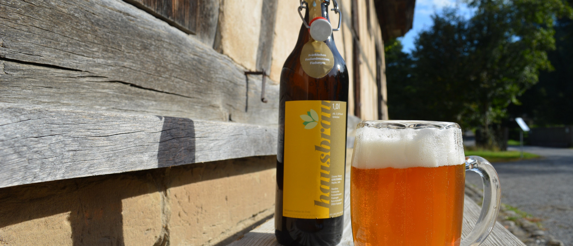 Bierflasche neben gefülltem Bierglas auf einer Holzbank vor Fachwerkhaus