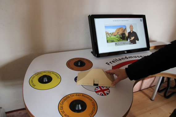 Tisch mit Bildschirm und verschiedenen 3D-Scannern. Man sieht einen Arm, der ein Hausmodell auf einen Scanner setzt.