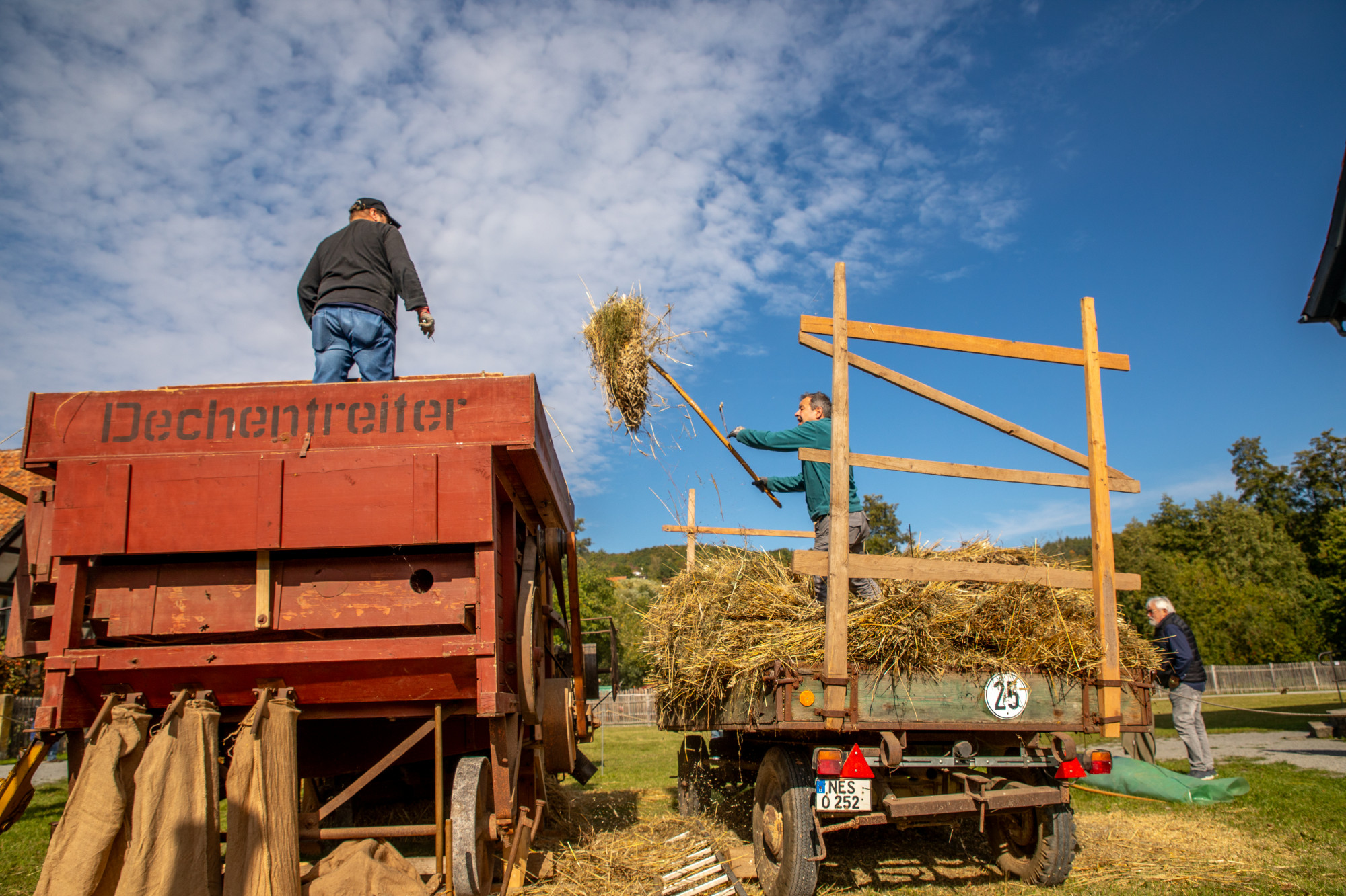 Zwei M&auml;nner laden Stroh von einem Anh&auml;nger auf eine rote Dreschmaschine mit der Beschriftung "Dechentreiter"