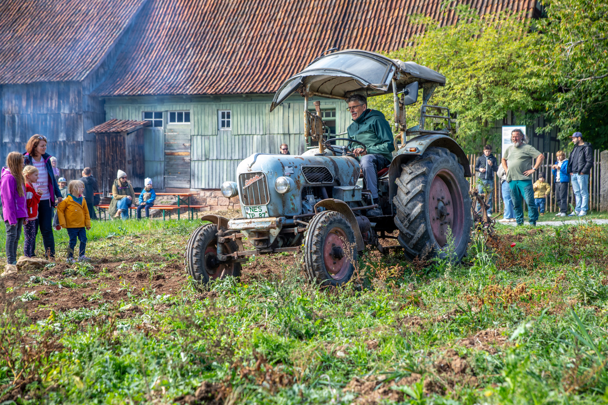 Mann bef&auml;hrt ein Feld auf einem Traktor, Personen stehen drumherum, , im Hintergrund historisches Geb&auml;ude und Baum