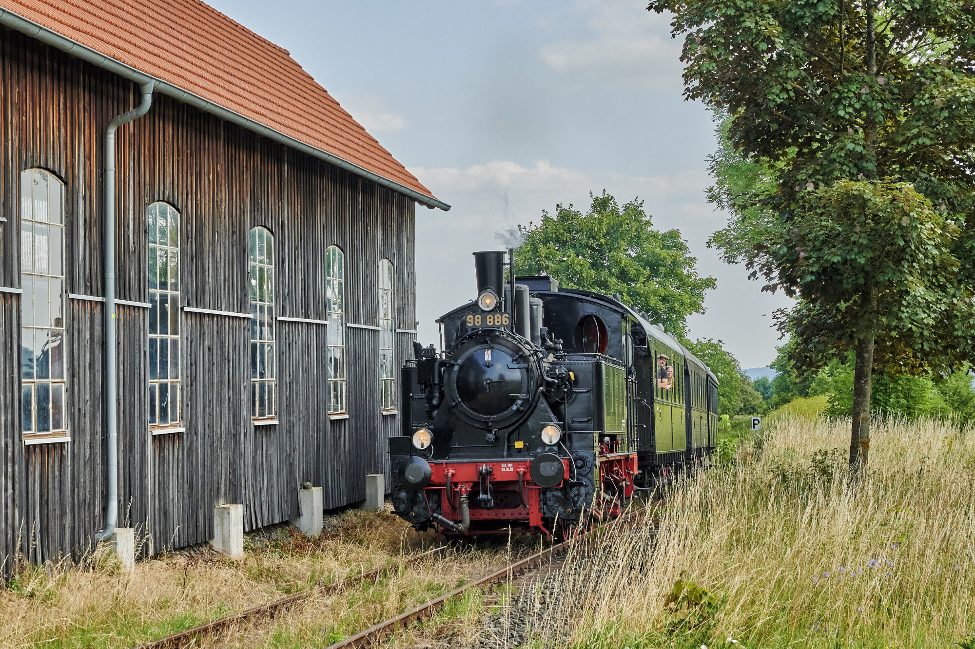 Dampfeisenbahn f&auml;hrt zwischen B&auml;umen und Lokschuppen entlang