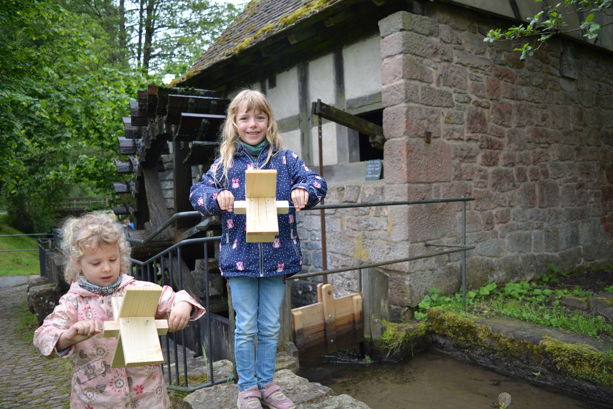 Zwei M&auml;dchen stehen vor einer M&uuml;hle und halten kleines Wasserrad aus Holz in der Hand