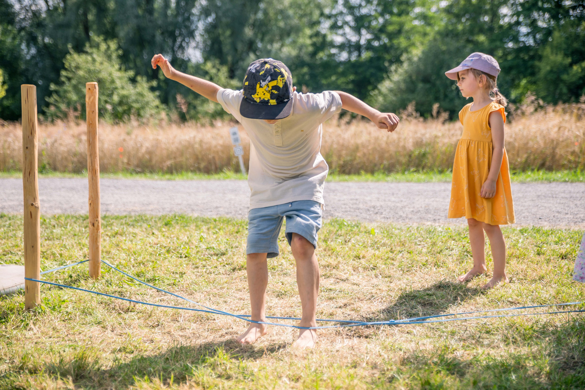 Zwei Kinder an einem Gummitwist, das an Holzpfl&ouml;cken befestigt ist, im Hintergrund Getreidefeld und B&auml;ume 