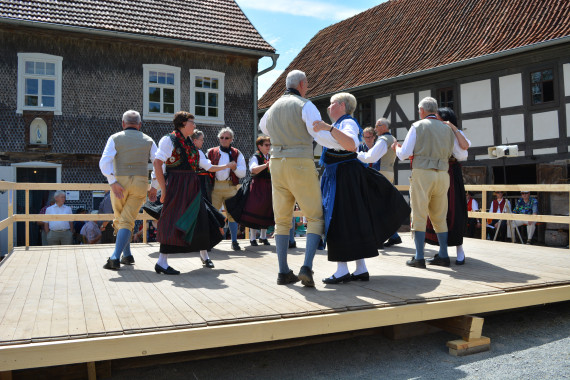 Mehrere Paare tanzen in Tracht auf einer B&uuml;hne vor historischen H&auml;usern