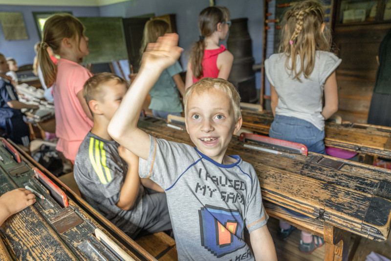 Kind mit grauem T-Shirt hebt in historischem Klassenzimmer die Hand, andere Kinder sitzen an alten Holzbänken