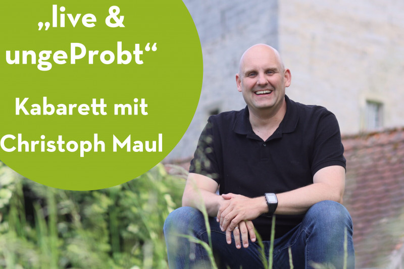 Mann in schwarzem Shirt sitzt vor Bäumen und historischem Gebäude