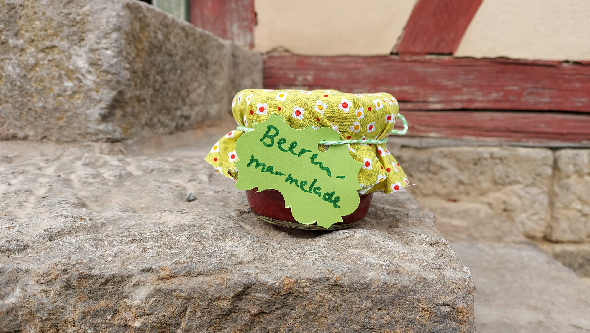 Ein Glas mit gr&uuml;nem Stoff&uuml;berzug und Etikett mit der Beschriftung "Beerenmarmelade" steht auf einer Steintreppe vor einer Fachwerkwand
