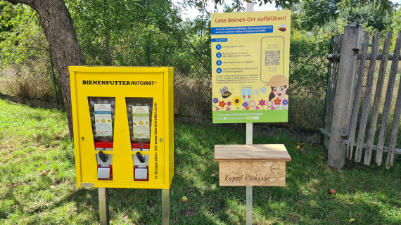 links ein gelber Bienenfutterautomat, rechts ein Hinweisschild, darunter eine Holzbox mit der Aufschrift "Kapselrückgabe"