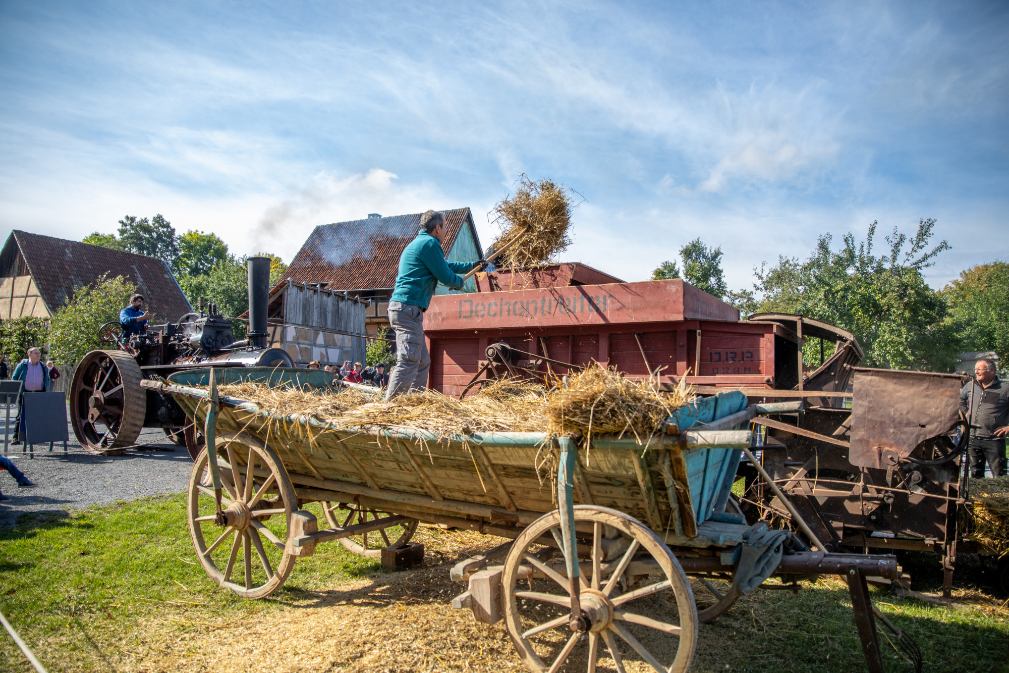 Person wirft Getreide auf eine historische Dreschmaschine im Freilandmuseum neben einem mit Stroh beladenen h&ouml;lzernen Wagen