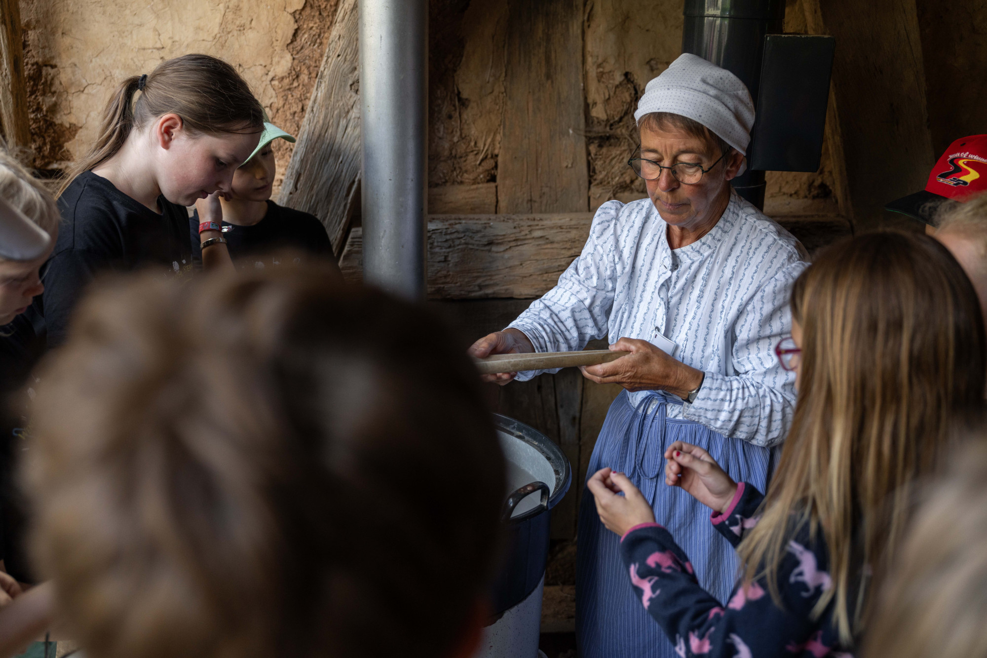 &Auml;ltere Frau in historischer Kleidung zeigt einer Gruppe von Kindern in einem rustikalen Innenraum eine traditionelle Waschtechnik.