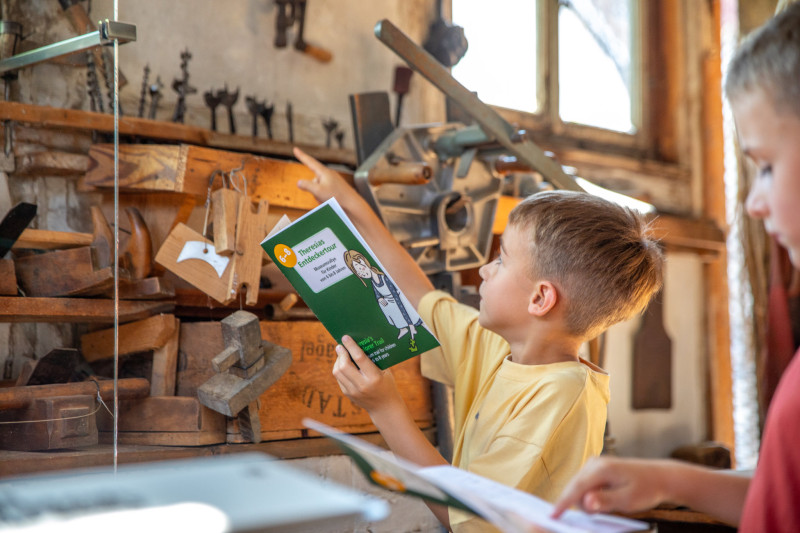 Junge in gelbem T-Shirt hält ein grünes Rallyheft und zeigt auf ein hölzernes Modell in einem Museum, im Vordergrund ein weiteres Kind mit Rallyheft